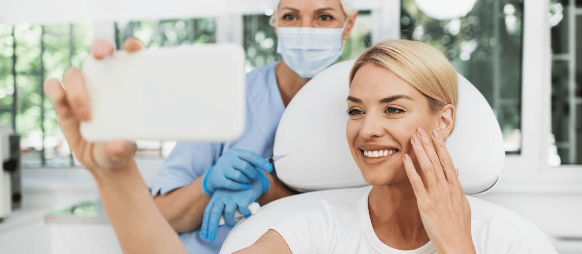 A dermatology provider smiling behind her patient while they look at her glowing skin in a handheld mirror.
